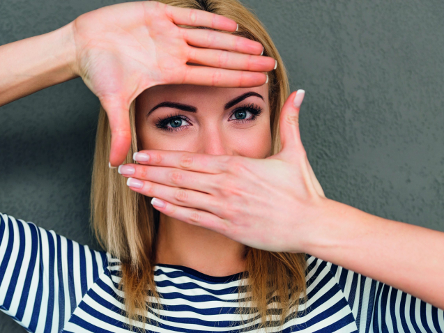Beautiful young woman making finger frame and looking at camera while standing against grey background